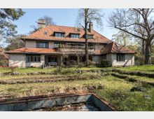 Großes Wohnhaus mit roten Ziegeldach und heller Fassade, umgeben von einem weitläufigen Garten mit Bäumen und Rasenflächen. Im Vordergrund sind gestufte Beete und ein betoniertes Wasserbecken sichtbar.