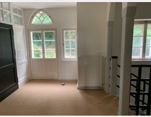 Ein heller Innenraum mit Teppichboden, Doppeltüren mit Sprossenfenstern und einem kleinen, halbkreisförmigen Fenster darüber. Neben den Türen ist ein Heizkörper, und rechts führt ein Treppengeländer aus Metall nach unten, flankiert von weißen Säulen.