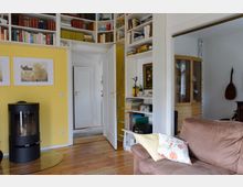 Wohnzimmer mit Holzboden, gelber Wand und eingebautem Bücherregal, das bis zur Decke reicht. Im Raum befinden sich ein braunes Sofa, ein Kaminofen, ein Glastisch sowie Türen, die in angrenzende Räume führen.