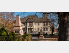 Ein historisches Gebäude mit heller Fassade und einem Walmdach, umgeben von Bäumen im Herbst. Im Vordergrund stehen zwei steinerne Denkmäler mit Inschriften und Sternsymbolen, eingebettet in einen kleinen Grünbereich.