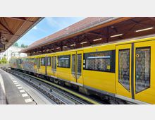 Gelber U-Bahn-Zug an einem erhöhten Bahnsteig mit überdachter Holzkonstruktion; im Hintergrund sind Wohngebäude und Bäume sichtbar. Der Bahnsteig ist mit Markierungen und einem geschwungenen Gleisverlauf gestaltet.