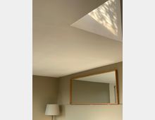 Minimalistisches Zimmer mit beiger Wand, einem großen rechteckigen Spiegel mit Holzrahmen sowie einer weißen Stehlampe. Tageslicht fällt durch ein dreieckiges Dachfenster, wodurch Licht- und Schattenspiele an der Decke sichtbar werden.