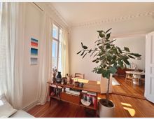 Helles Wohnzimmer mit Holzdielen, einer Zimmerpflanze in einem großen Topf und einem Holztisch mit Büchern und Dekoobjekten. Im Hintergrund sind ein Esstisch und weitere Möbel sichtbar, das Licht fällt durch hohe Fenster mit weißen Vorhängen.