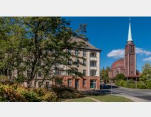 Eine Straßenansicht mit einem beige-braunen Wohnhaus links, das von Bäumen teilweise verdeckt ist, sowie einer roten Backsteinkirche mit Kuppel und spitzem Turm rechts im Bild. Der Vordergrund zeigt eine Grünfläche und eine Kreuzung unter klarem, blauem Himmel.