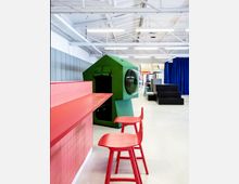 Ein moderner Innenraum mit einer Kombination aus bunten Möbeln: eine rote Theke mit rosafarbenen Fliesen und zwei roten Stühlen im Vordergrund, sowie eine grüne, hausförmige Sitzbox mit einem runden Fenster dahinter. Der Raum hat weiße Wände, eine offene Decke mit sichtbaren Leitungen und hängt mit blauen Vorhängen dekorativ ab.