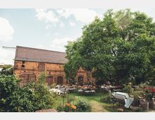 Ein rustikaler Hof mit einer Backsteinscheune im Hintergrund, umgeben von grünem Laub und Bäumen. Im Vordergrund stehen zahlreiche Tische und Stühle auf einer Wiese, die wie ein Außenbereich einer Veranstaltung oder eines Cafés wirken, umrahmt von Pflanzen und dekorativen Elementen.