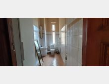 Schmales, längliches Badezimmer mit hohen Decken, weiß gefliesten Wänden mit Zierbordüre und braunem Bodenbelag. Am Ende des Raums stehen WC und ein hohes, schmales Fenster; seitlich sind ein kleines Waschbecken, ein Heizkörper und ein an die Wand gelehnter Spiegel zu sehen.
