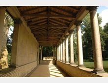 Überdachter Säulengang mit klassizistischen Elementen, bestehend aus korinthischen Säulen und einem Holzsatteldach, angrenzend an eine Steinwand; im Hintergrund ein Park mit Bäumen und Wiese.