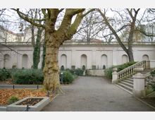 Ein Park mit einem großen Baum im Vordergrund, herbstlichem Laub auf dem Boden und einer breiten Treppe mit steinernen Geländern rechts. Im Hintergrund ist eine helle historische Mauer mit Arkadenbögen zu sehen, umgeben von Büschen und kletternden Pflanzen.