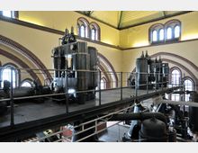 Ein industrieller Innenraum mit historischen Maschinen, darunter metallene Zylinder und Rohrkonstruktionen, auf einer erhöhten Plattform. Die Wände sind gelb getüncht und mit roten Backsteinbögen verziert, die hohe Fenster mit Rundbögen umrahmen.