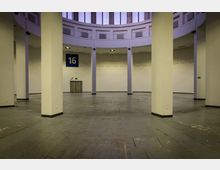 Runder, großflächiger Innenraum mit hohen weißen Säulen und einer offenen Decke, durch die Fensterreihen Licht einfallen. Eine blaue Tafel mit der Nummer „16“ hängt an der Wand, die von weißen Paneelen und einer Tür geprägt ist.