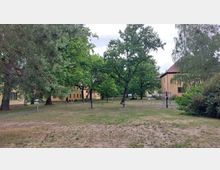 Ein parkähnlicher Bereich mit Rasenfläche, vereinzelten Bäumen und gelbgestrichenen Gebäuden mit roten Dächern im Hintergrund. Im Vordergrund befinden sich mehrere Fitnessgeräte im Freien sowie ein gepflasterter Weg.