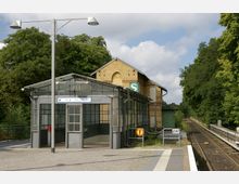 Eingangsbereich eines kleinen S-Bahnhofs mit Glasfassade und Ziegeldach, flankiert von einem gelben Backsteingebäude. Rechts im Bild verläuft das Gleis neben einer Baumreihe.