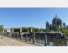 Flusslandschaft mit Brücke im Vordergrund, geschmückt mit Skulpturen, und dem Berliner Dom im Hintergrund, gekrönt von grünen Kuppeln. Rechts verläuft die Uferpromenade mit Bäumen und einer U-Bahn-Station, links ist ein gepflasterter Gehweg mit Laternen.