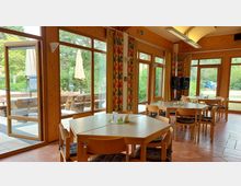 Ein heller Gemeinschaftsraum mit großen Glasfenstern, durch die eine Terrasse mit Holzbänken und Bäumen sichtbar ist. Holzverkleidete Wände, gemusterte Vorhänge, und einfache Holztische mit Stühlen prägen die Einrichtung; die Tischdekoration besteht aus kleinen Karten und Geschirr.