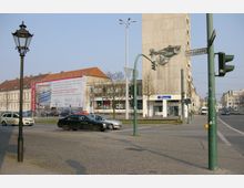Straßenszene an einer Kreuzung mit gepflastertem Gehweg, Laterne und Ampeln; ein Plattenbau mit einem Wandmosaik und ein historisches Gebäude im Hintergrund, teils verdeckt durch ein Baugerüst mit Werbeplakat. Verkehr mit geparkten und fahrenden Autos sichtbar.