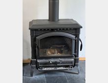 Ein schwarzer Holzofen aus Metall mit einer großen Glastür, einem Kaminrohr nach oben und einem Aschefach unten. Der Ofen steht auf einem dunklen Steinboden vor einer weißen Wand.