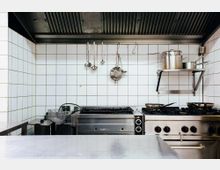 Professionelle Industrieküche mit weißen Fliesenwänden, Edelstahl-Herd und Dunstabzugshaube. Kochutensilien und Töpfe hängen an einer Stange oder stehen auf einem Regal.