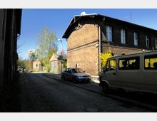 Eine gepflasterte Seitenstraße mit Ziegelgebäuden auf beiden Seiten, darunter ein zweistöckiges Haus mit weißen Fensterrahmen. Im Vordergrund stehen ein geparkter Kleintransporter und ein Auto, während im Hintergrund Bäume und weitere Gebäude sichtbar sind.