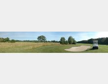 Weitläufiger Golfplatz mit gepflegten Rasenflächen, einem Sandbunker und hohen Gräsern im Hintergrund; ein Golfcart steht rechts neben dem Sandbunker. Im Hintergrund sind vereinzelte Bäume und eine lockere Baumreihe sichtbar.