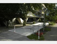 Mehrstöckiges Wohngebäude mit Balkonen, das von massiven, schrägen Pfeilern getragen wird. Vor dem Gebäude befindet sich ein barrierefreier Zugang mit einer Rampe, umgeben von Bäumen, Blumenbeeten und gepflasterten Wegen.