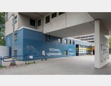 Modernes Stadtgebäude mit blauer Fassade, bedeckt mit Graffiti, unteren Fenstern und einem überhängenden Betonteil; im Vordergrund Gehweg und Geländer, rechts der Eingang zu einem Gebäudeteil.