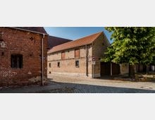Kopfsteingepflasterte Straßenecke mit zwei historischen Backsteingebäuden mit Satteldächern, kleinen vergitterten Fenstern und hölzernen Klappläden. Rechts steht ein großer Baum am Gehweg neben Einfahrtstoren und einem Verkehrsschild.