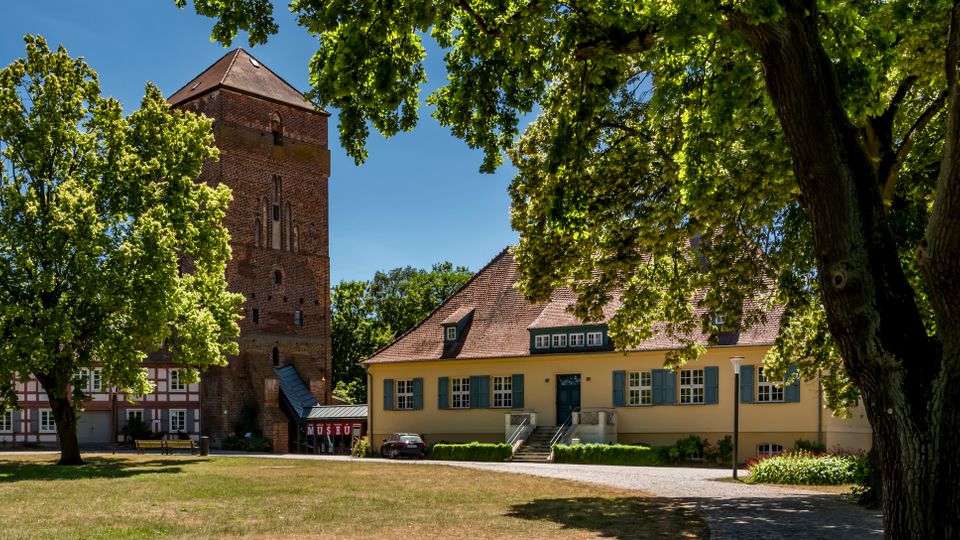 Blick auf einen historischen Hof mit großer Rasenfläche und alten Laubbäumen, im Hintergrund ein hoher Backstein-Turm sowie ein langes, gelb gestrichenes Gebäude mit rotem Ziegeldach und blauen Fensterläden. Links stehen weitere Fachwerkbauten, und ein Weg führt zum Eingang des Gebäudes.