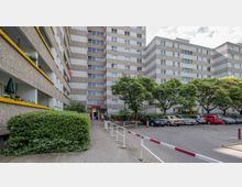 Mehrstöckiges Wohngebäude mit grauer Fassade und gelben Balkonbrüstungen, umgeben von Bäumen. Im Vordergrund befinden sich ein Parkplatz mit geparkten Autos und ein befestigter Gehweg mit Absperrungen.