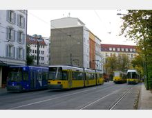 Straßenszene mit mehreren Straßenbahnen, darunter gelbe und eine blaue Straßenbahn, die auf parallelen Gleisen in einer städtischen Umgebung stehen. Im Hintergrund sind mehrstöckige Wohngebäude und Bäume zu sehen, Straßenlaternen und Oberleitungen flankieren die Gleise.