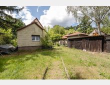 Hinterhof mit grasbewachsener Fläche und schmalem Weg zwischen einem kleinen Haus mit Satteldach und mehreren Holzschuppen. Umgeben von Bäumen und Sträuchern, mit Zäunen und Geräten an der Schuppenwand.