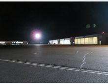 Flughafengelände bei Nacht mit beleuchteten Hangars im Hintergrund und einer großen, leeren Asphaltfläche im Vordergrund. Eine helle Lampe in der Mitte des Bildes strahlt Licht aus, während im linken Bereich kleine Flugzeuge vor den Hangars sichtbar sind.