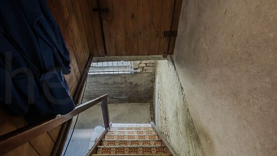 Blick in ein schmales, alt wirkendes Treppenhaus mit hölzerner Decke und abgenutzten Putzwänden. Eine steile Treppe mit gemustertem Belag und Holzgeländer führt hinunter zu einem Kellerbereich mit freiliegendem Mauerwerk und kleinem Fenster.