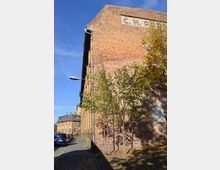 Backsteinfassade eines historischen Industriegebäudes mit der Aufschrift „C.H. P...“. Im Vordergrund stehen mehrere junge Bäume, rechts davon fällt Schatten, und links ist ein Gehweg mit parkenden Autos zu sehen. Der Himmel ist klar und blau.