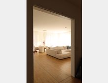 Blick in ein modernes Wohnzimmer mit einem hellen Holzfußboden, einem großen cremefarbenen Ecksofa und einem runden Esstisch mit Stühlen in einer Ecke. Die Räume sind durch große Fenster mit weißen Vorhängen gut beleuchtet.