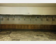 Ein leerer Garderobenbereich mit einer langen Holzablage und mehreren Metallhaken für Kleiderbügel an einer hellen Wand, beschriftet mit "1–200". Der Boden ist mit grauen und weißen Marmorkacheln gefliest.