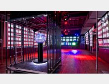 Ein moderner Clubraum mit industriellem Design, Holzboden und einer zentralen Lichtinstallation. Die Wände und Decke sind mit leuchtenden LED-Displays dekoriert, die Zahlenmuster zeigen, und der Raum ist in blaues und rotes Licht getaucht.