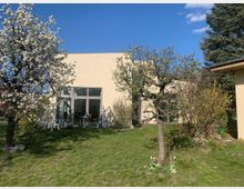 Ein moderner, flacher Gebäudekomplex mit beiger Fassade und großen Glasfenstern steht inmitten eines grünen Gartens mit blühenden Bäumen und Sträuchern. Ein blauer Himmel und vereinzelte Wolken bilden den Hintergrund.