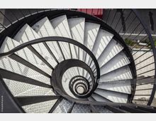 Spiralförmige Metalltreppe mit rutschfesten, diamantgemusterten Stufen, die von einem schwarzen Geländer umgeben ist und in einer Ansicht von oben fotografiert wurde. Im Hintergrund ist ein Außenbereich mit Gehweg und Straße sichtbar.