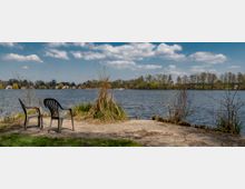 Zwei Plastikstühle stehen auf einer befestigten Fläche am Ufer eines Sees, umgeben von Gras und Sträuchern. Im Hintergrund sind das Wasser, eine Baumreihe und vereinzelte Häuser auf der gegenüberliegenden Uferseite zu sehen, darüber ein blauer Himmel mit weißen Wolken.