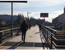 Fußgängerbrücke mit Geländern auf beiden Seiten, die über ein urbanes Gebiet mit modernen Gebäuden führt. Im Hintergrund ist ein Werbebildschirm zu sehen, und mehrere Personen gehen auf der Brücke bei sonnigem Wetter.