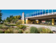 Eine erhöhte Betonbrücke mit blauen Glasgeländern und gelb gestrichenen Pfeilern, unter der ein Gehweg und Begrünung verlaufen. Im Vordergrund befinden sich bepflanzte Beete mit Gräsern und Blumen, umgeben von Bäumen und Sträuchern.