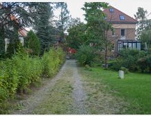 Eine schmale, mit Kies bedeckte Zufahrtsstraße führt durch einen Gartenbereich mit Rasenflächen, Büschen und Bäumen. Im Hintergrund stehen mehrere Wohnhäuser mit roten Ziegeldächern und teilweise verglasten Anbauten.