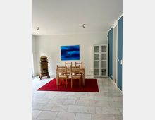 Helles Esszimmer mit weiß gefliesten Bodenplatten, einem Holztisch mit sechs Stühlen auf einem roten Teppich und einem großen blau abstrakten Wandbild. Rechts steht ein weißer Glasvitrinenschrank neben einer blau akzentuierten Wand, links ein dekorativer antiker Ofen.