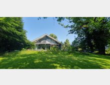 Modernes Einfamilienhaus mit Satteldach und großen Fensterfronten, umgeben von einem weitläufigen, grünen Garten mit Bäumen im Hintergrund. Der Rasen ist von Schatten und Sonnenlicht durchzogen, und die Umgebung wirkt naturbelassen.