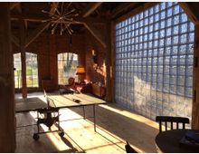 Ein Innenraum mit rustikaler Ziegelbauweise, großen rechteckigen Fenstern und einer Wand aus Glasbausteinen, die Tageslicht einlässt. In der Mitte steht eine Tischtennisplatte, daneben befinden sich eine Sitzecke mit roten Möbeln und eine Stehlampe.