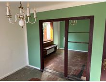 Ein einfacher Innenraum mit Holzdielenboden, grün gestrichenen Wänden und einer dunklen Holztüröffnung, die in einen weiteren Raum führt. Eine weiße Deckenleuchte mit mehreren Armen hängt von der Decke, und ein Fenster mit Holzrahmen lässt natürliches Licht herein.