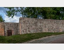 Eine historische Steinmauer aus unregelmäßigem Naturstein und Ziegeln, mit einem kleinen Holztor links. Im Hintergrund sind Bäume und ein teilweise bewölkter Himmel zu sehen, während im Vordergrund eine gepflasterte Fläche und Gras erkennbar sind.