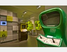 Ein Ausstellungsraum mit Retro-Computerspielen und Konsolen, darunter ein grüner Arcade-Automat im Vordergrund und ein "Pong"-Spielautomat in der Mitte. Die Wände sind mit weißen und limonengrünen Regalen sowie Informationstafeln dekoriert.