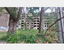 Ein verlassenes, mehrstöckiges Gebäude mit zahlreichen offenen Fensteröffnungen, umgeben von dichter Vegetation und hohen Bäumen. Im Vordergrund sind Graffiti auf Mauern und ein Zaun an der rechten Bildseite sichtbar.